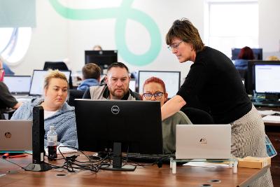 Three people are sat looking at a computer. A woman stood next to them showing them something on the screen. 