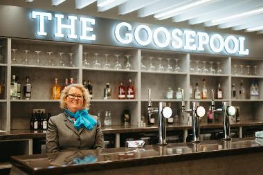 A woman wearing a uniform standing in front of a bar. There is a sign that reads "The Goosepool" above her head.