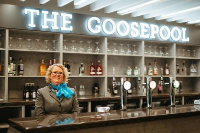 A woman wearing a uniform standing in front of a bar. There is a sign that reads "The Goosepool" above her head.