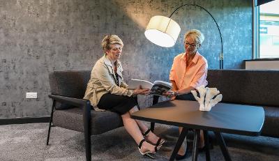 Two smiling women sat around a table at Lusso look at a brochure, brightly lit by a floor lamp